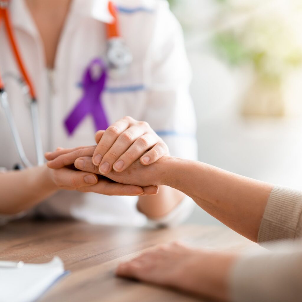 Blog 3 Close-up image of a doctor's hand, wearing a purple ribbon for cancer awareness, gently holding a patient's hand. conveys empathy and support,