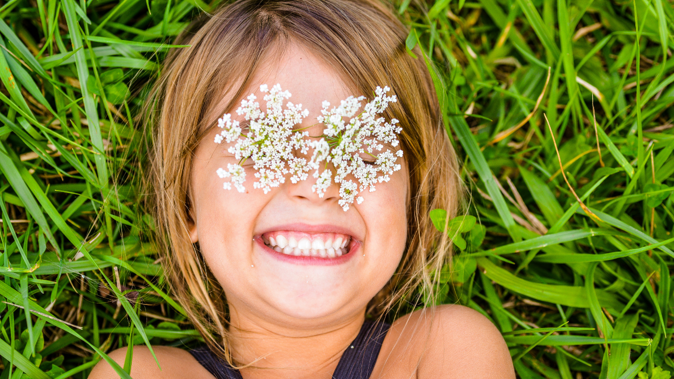 A young girl smiles brightly lying in a grassy garden, with baby's breath flowers on her eyes.