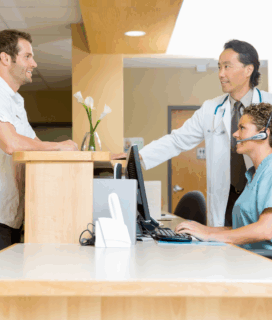 A smiling doctor and nurse at Southwest Urgent Care in Sugar Land, TX, warmly assisting a Telfair Plaza employee at the reception desk, showcasing community care and friendly service.